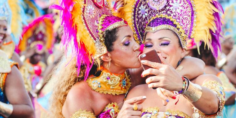 Two women Carnival dancers pose for a selfie as one kisses other on the cheek.